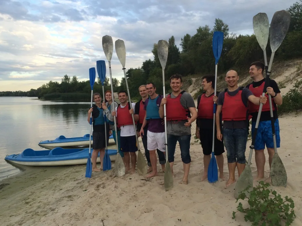 A group of people wearing life jackets hold paddles on a sandy beach next to blue kayaks and a river.
