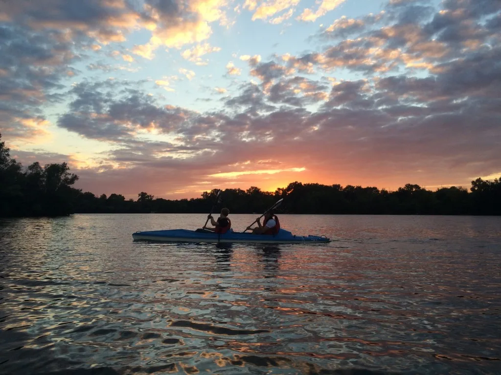 Two people in a blue kayak on a river at sunset.