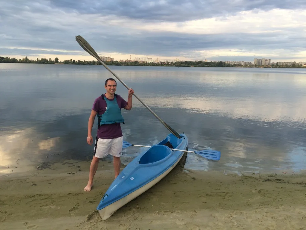 A man wearing a life vest stands on a sandy beach next to a blue kayak and holds a paddle.
