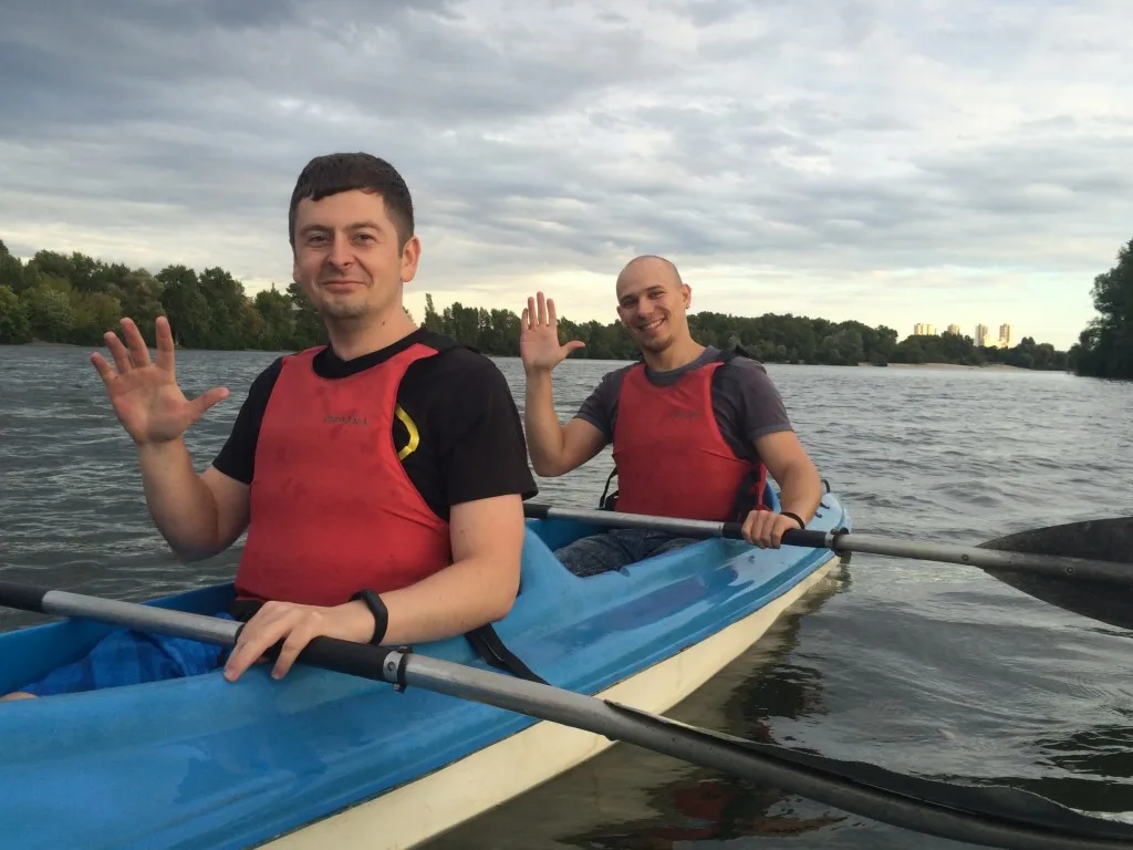 Two men wearing red life vests wave while sitting in a blue rowing boat on a river.