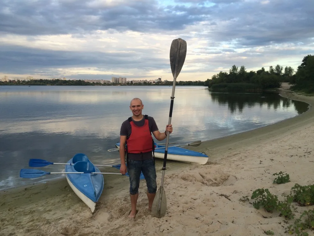 A man wearing a red life vest stands on a sandy beach with a paddle and two kayaks.